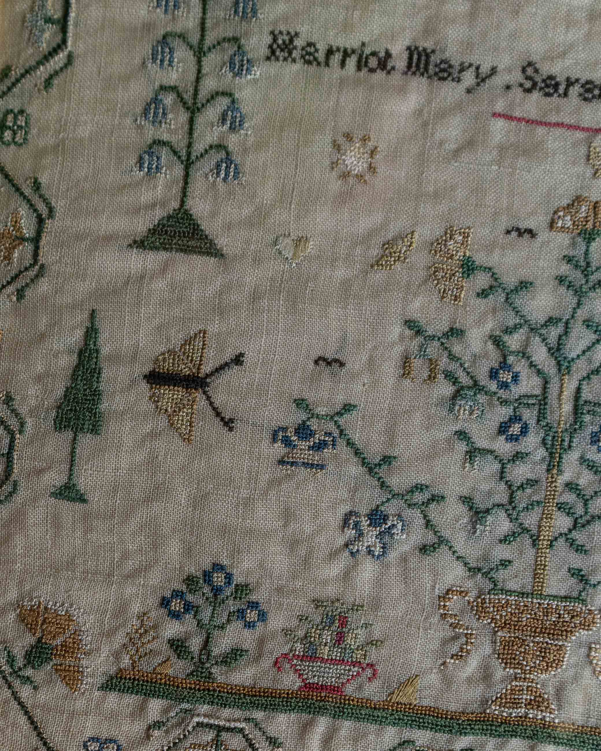 Embroidered sampler with floral patterns, trees, and butterflies on a beige background.