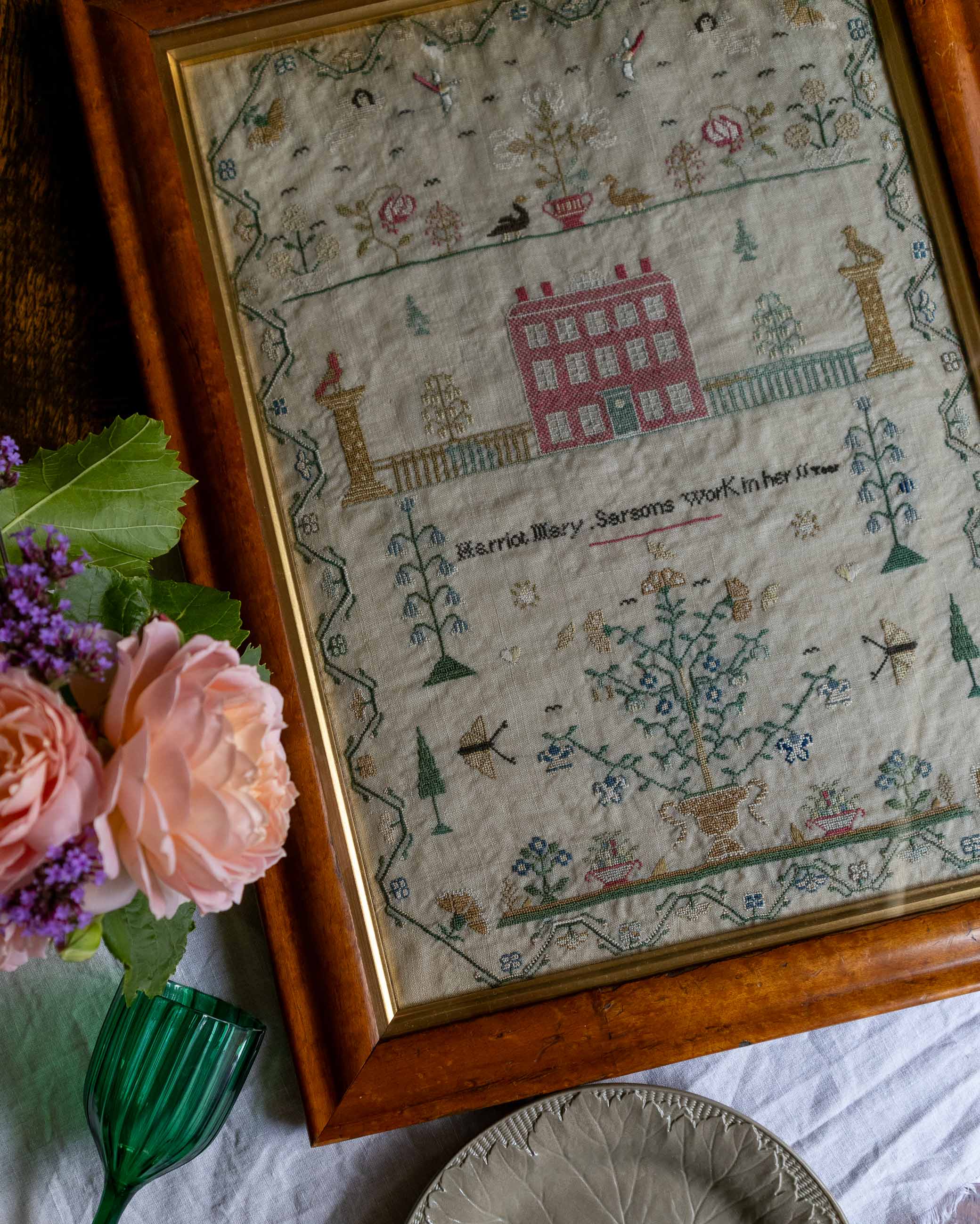 Framed embroidered sampler with floral decorations and text, placed on a surface with flowers and a vase.