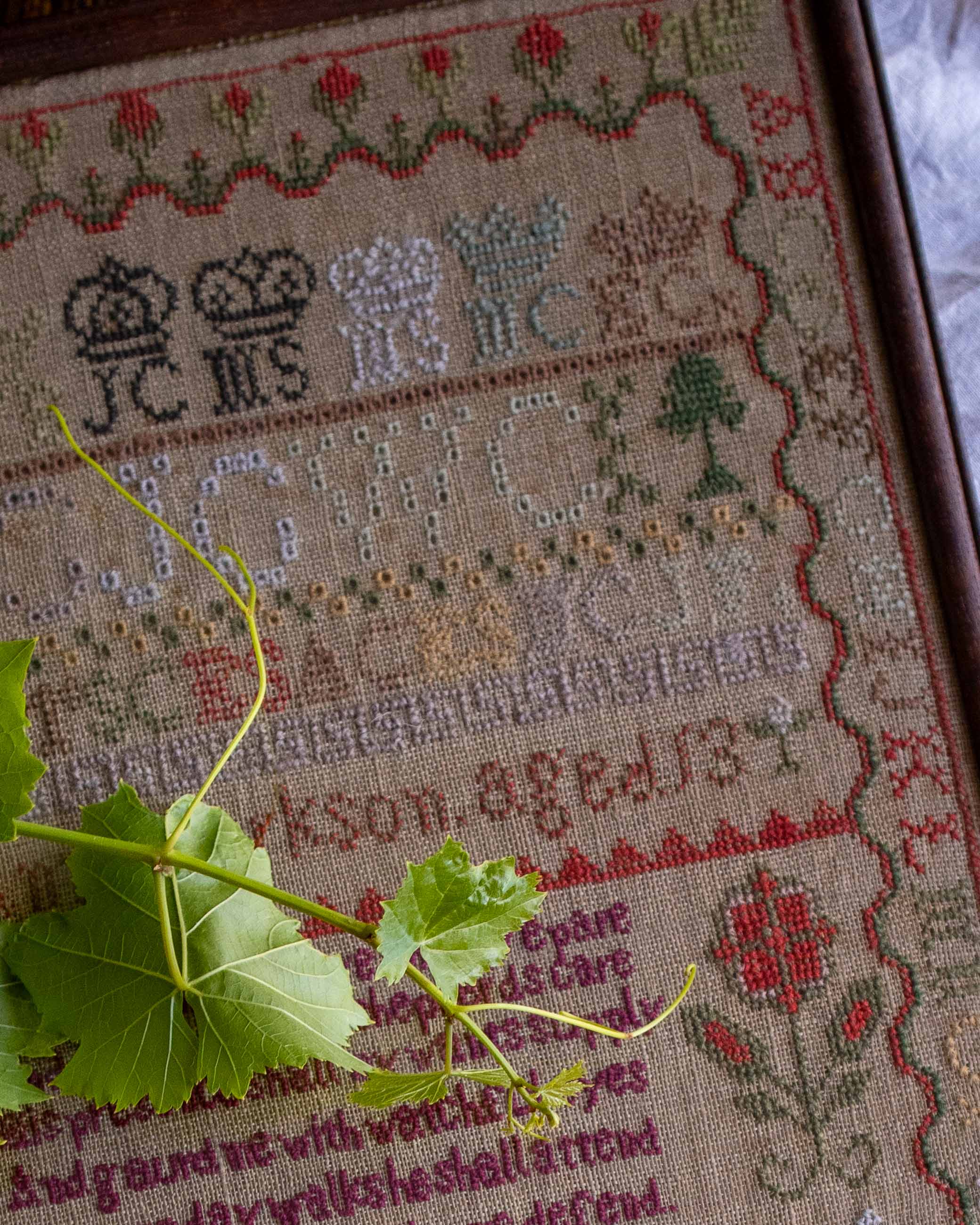 Close-up of a embroidered sampler with text and symbols, featuring a green leaf in the foreground.