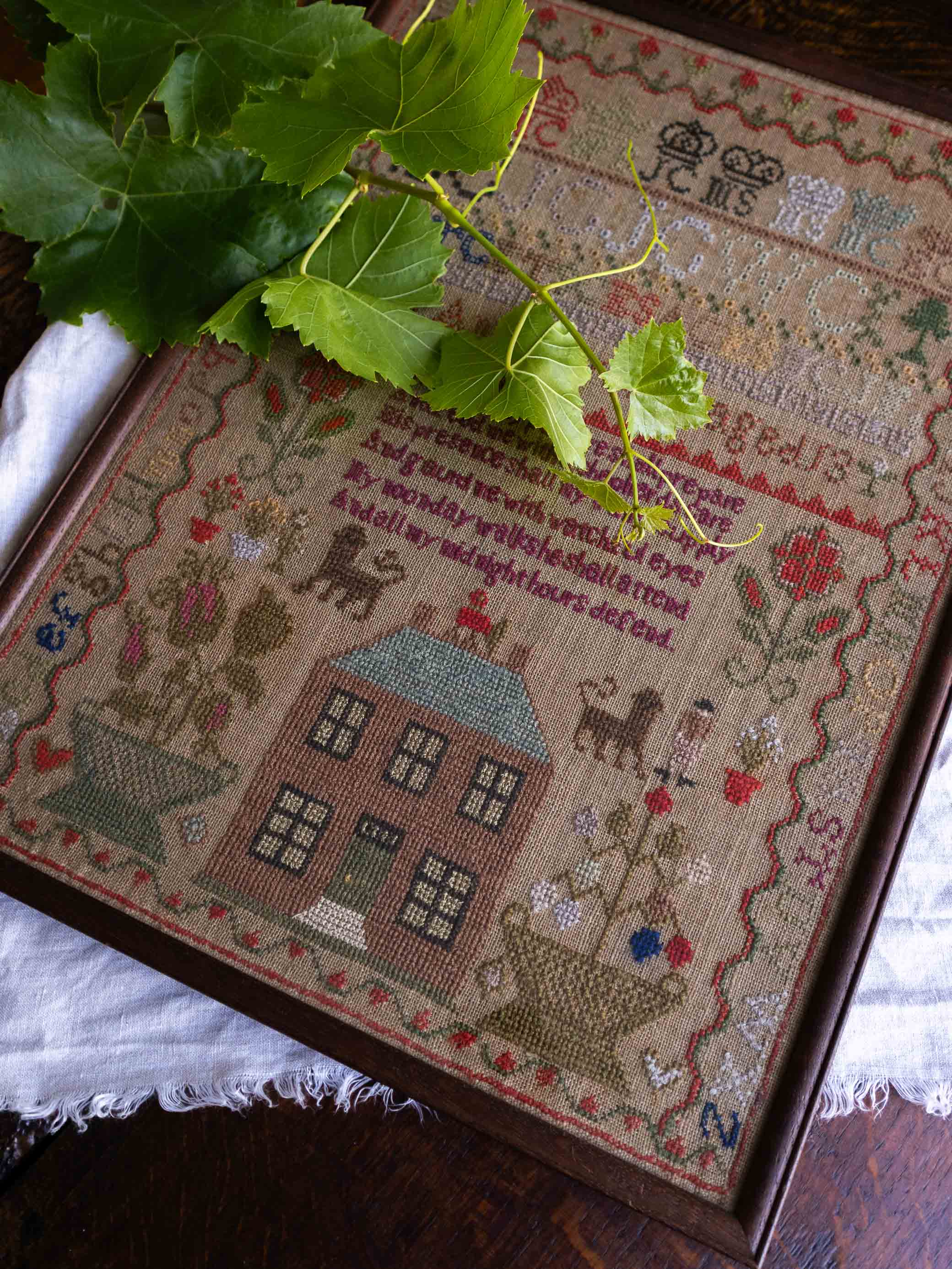 Framed cross-stitched antique sampler with a house design and decorative elements on a wooden surface.