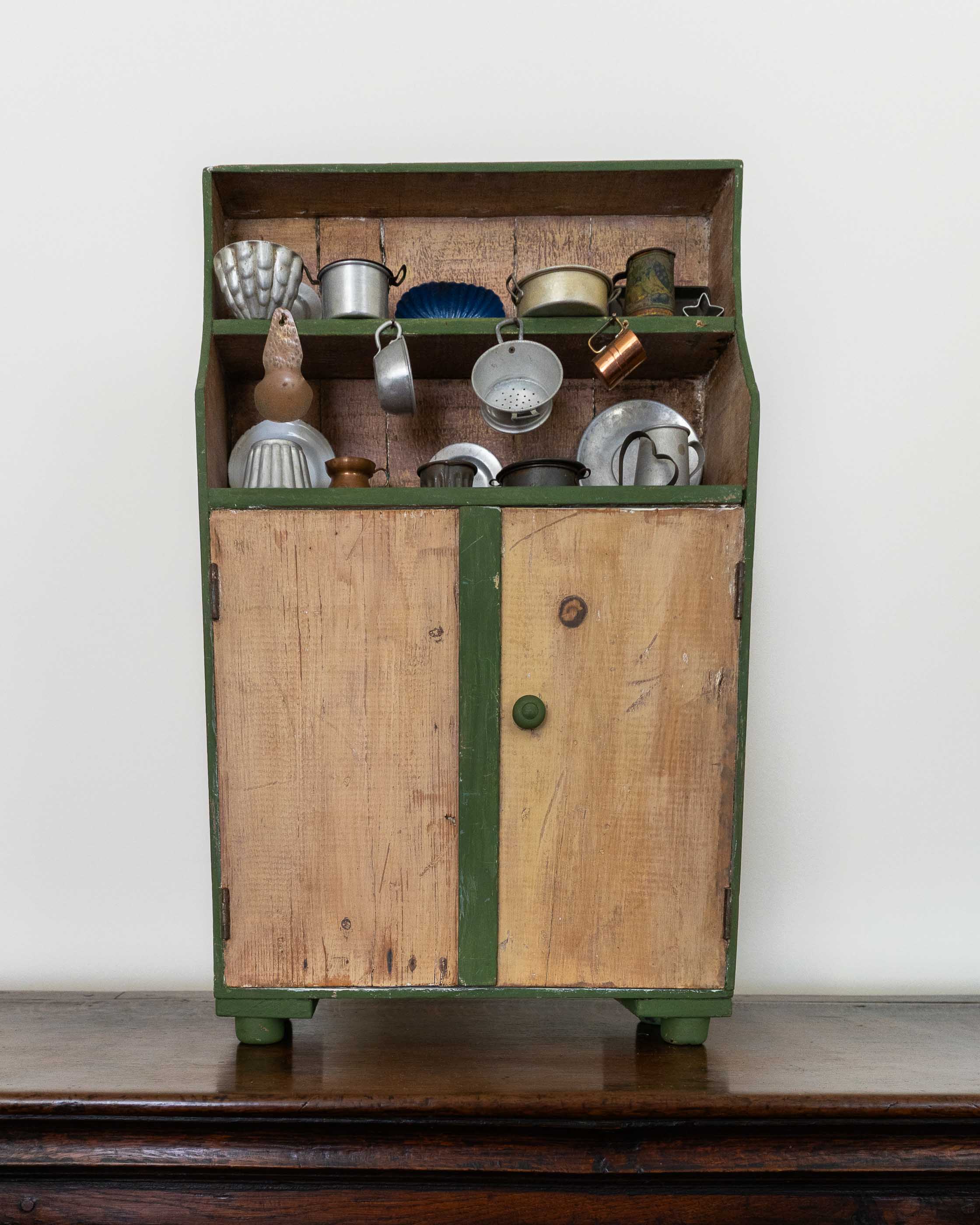 Vintage wooden toy cabinet with green trim filled with metallic kitchenware on a wooden surface.