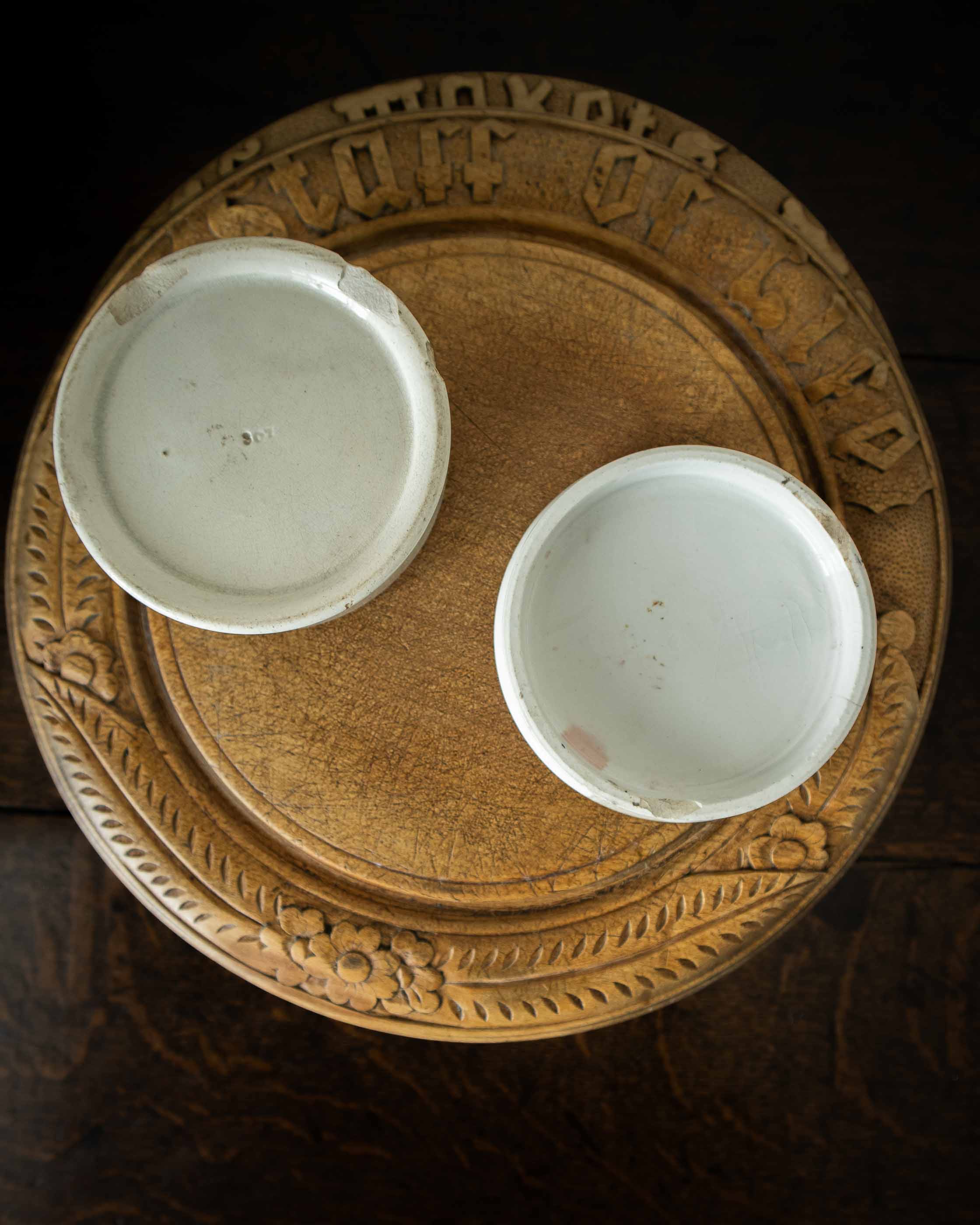 White ceramic pot with lid on a wooden breadboard with intricate carvings.