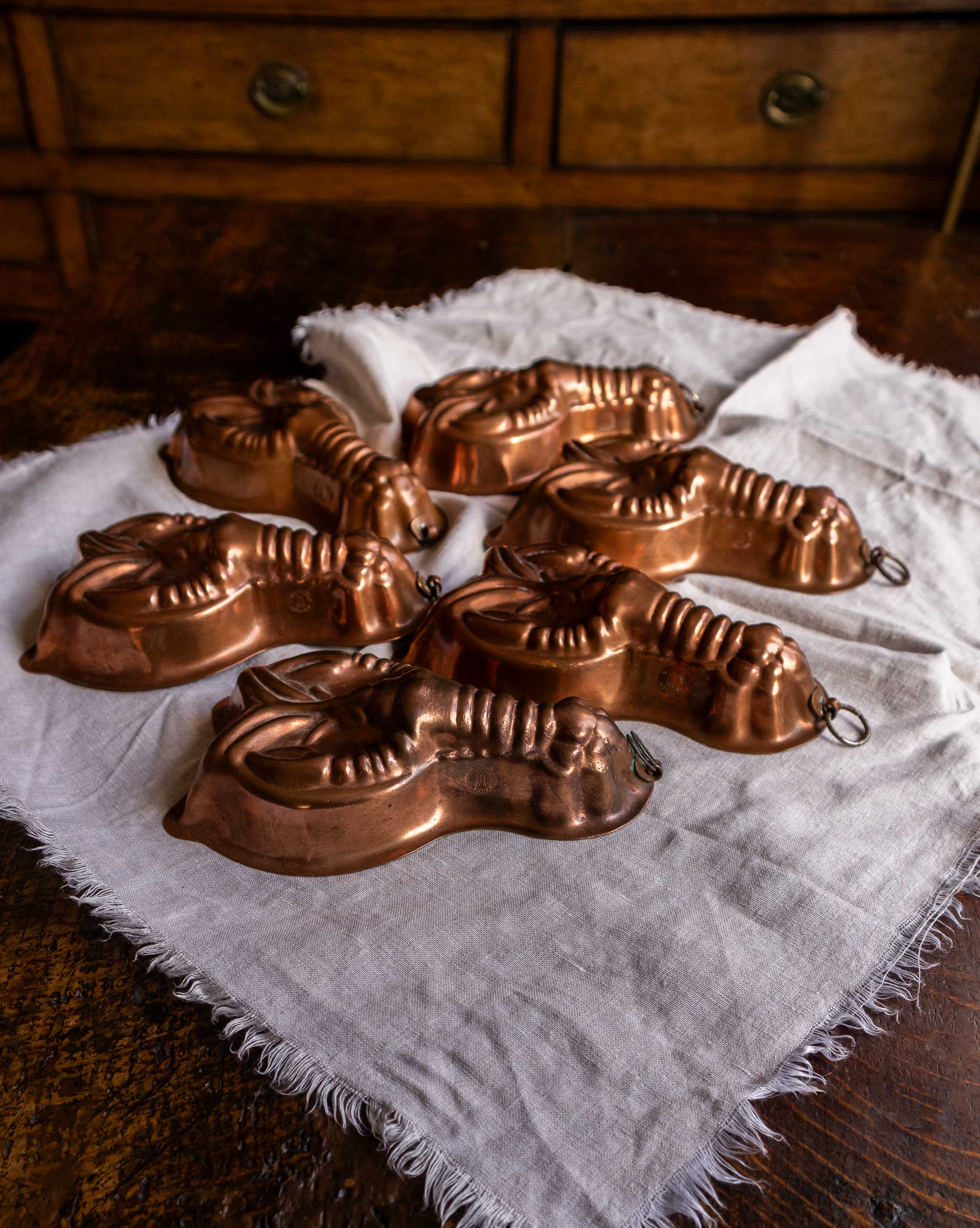 Set of copper lobster-shaped molds on a white cloth with wooden drawers in the background.