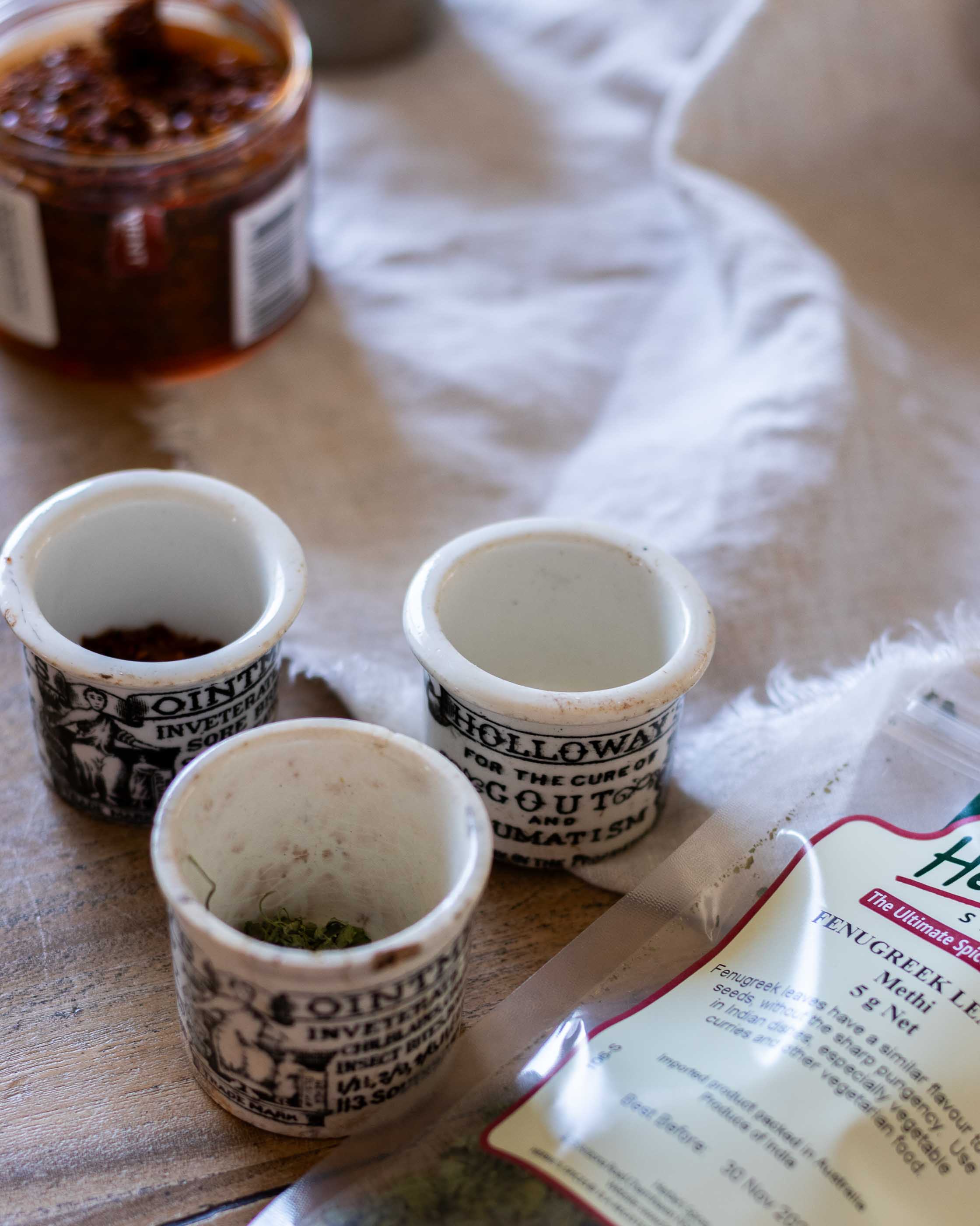 Three small ceramic pots with text on a wooden surface next to a jar and a packet of herbs.
