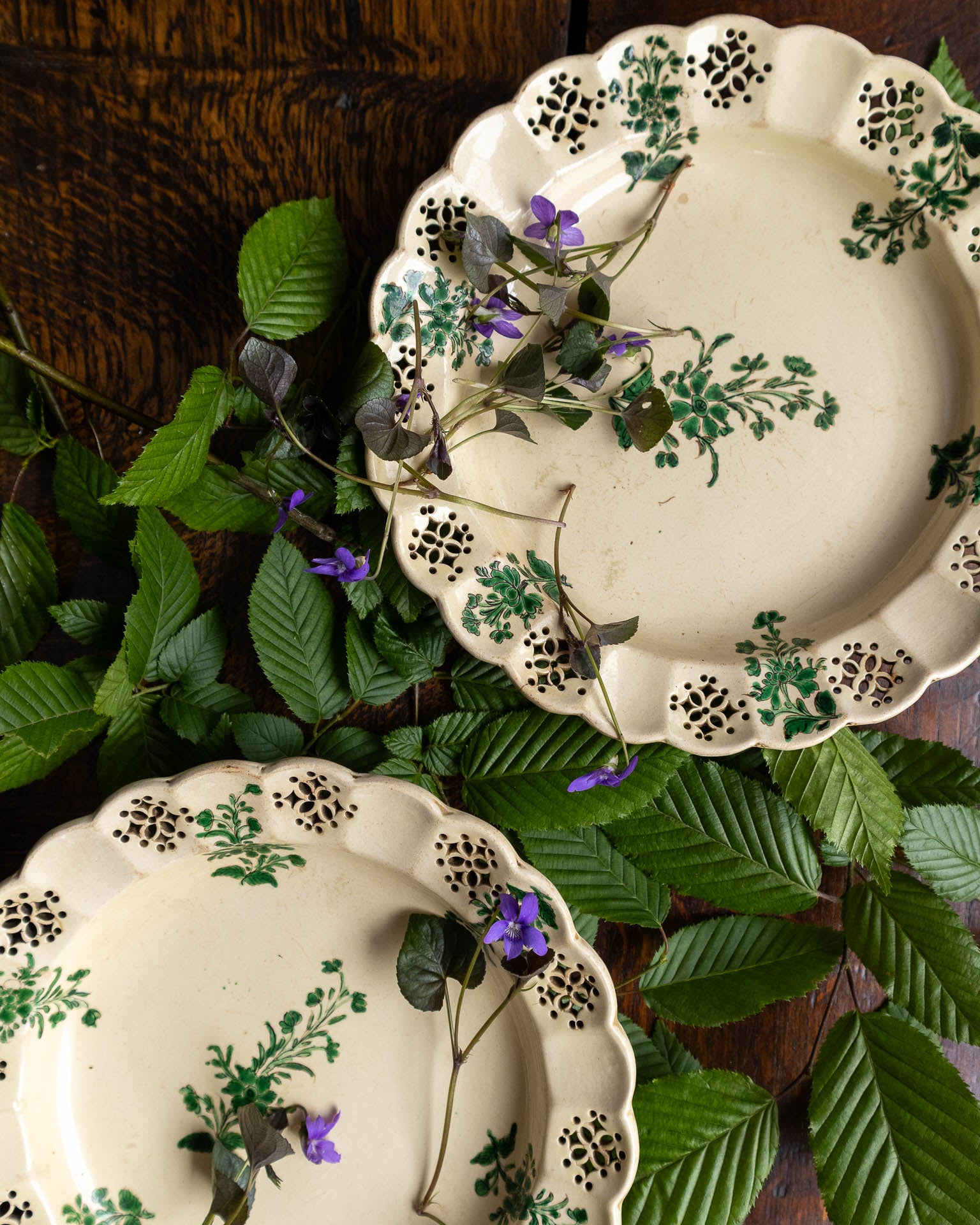 Two decorative creamware plates with floral patterns on a bed of green leaves.