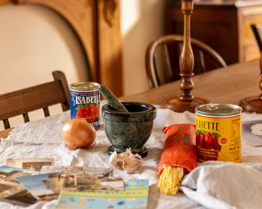 A table laid with pasta, cookbook, holiday images, tins of tomatoes and a mortar and pestle.