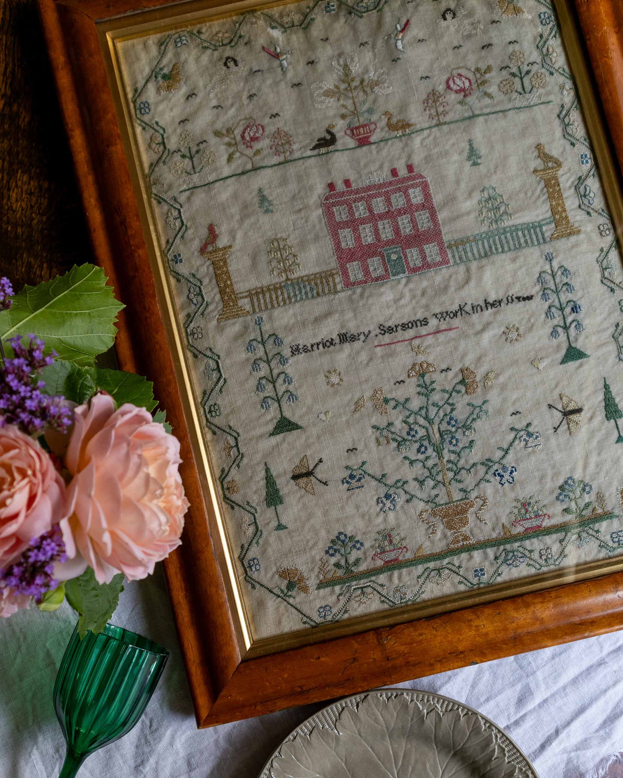 Framed embroidered sampler with floral decorations and text, placed on a surface with flowers and a vase.