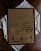 Framed antique cross-stitched sampler on a wooden surface with a white cloth underneath