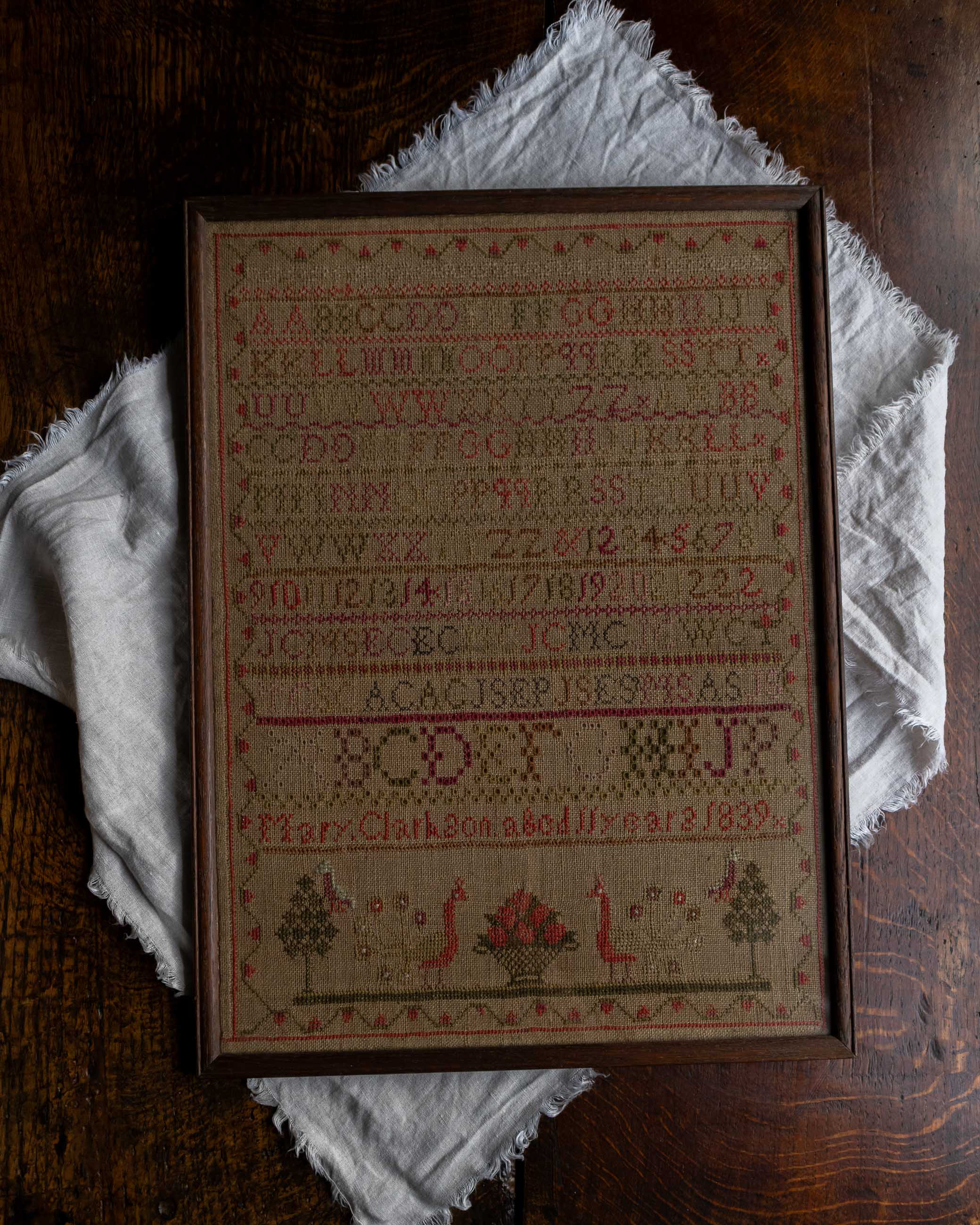 Framed antique cross-stitched sampler on a wooden surface with a white cloth underneath