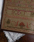 Close-up of a framed embroidered sampler with text and designs on a wooden surface.