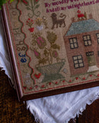 Close-up of a cross stitch antique sampler with a house and cat design on a textured surface.