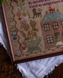 Close-up of a cross stitch antique sampler with a house and cat design on a textured surface.