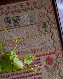 Close-up of a embroidered sampler with text and symbols, featuring a green leaf in the foreground.