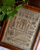 Framed cross stitch sampler with intricate designs and text, placed on a surface with green leaves.