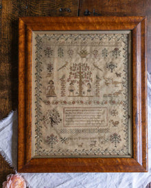Framed sampler with intricate patterns including Adam and Eve and text on a wooden surface