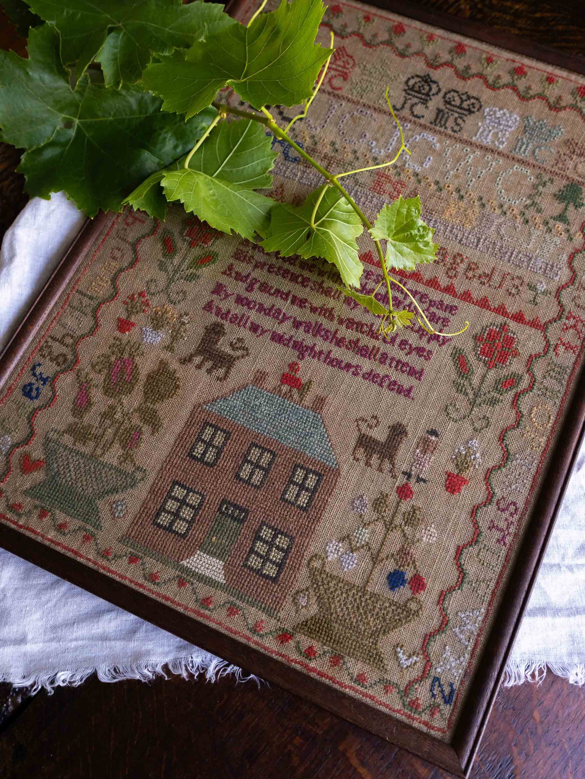 Framed cross-stitched antique sampler with a house design and decorative elements on a wooden surface.