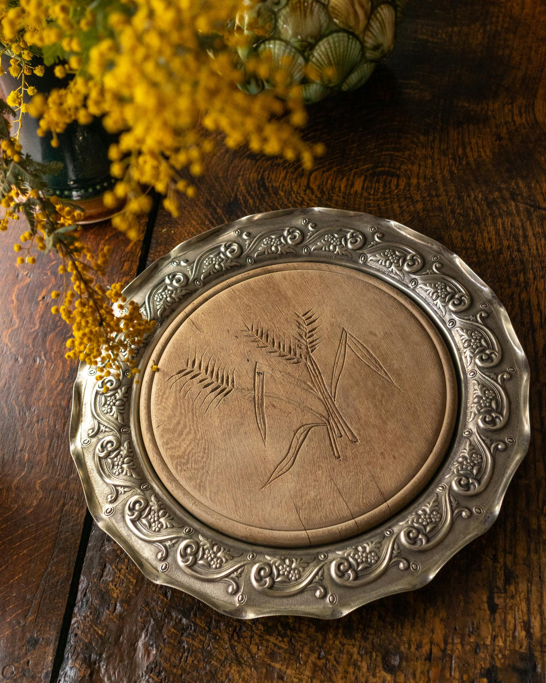 Decorative silver plated breadboard with carved design on a wooden surface with yellow wattle flowers.