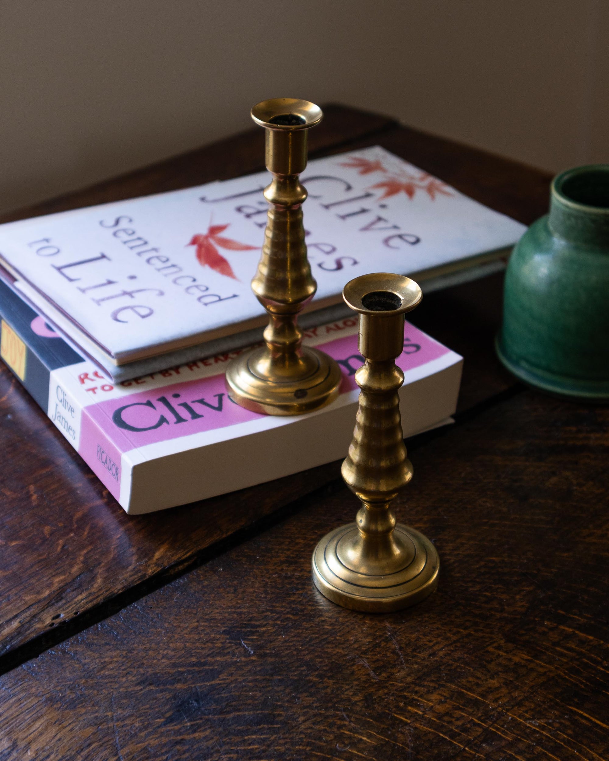 Two brass candlesticks on a wooden surface with books and a green vase in the background.