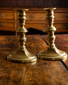 Two brass candlesticks on a wooden surface with a wooden drawer in the background.