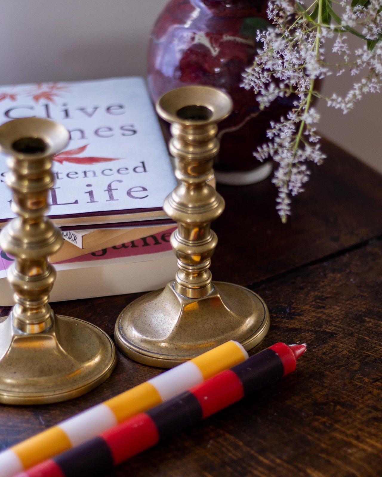 Brass candlesticks with colorful candles on a wooden surface next to books and flowers.