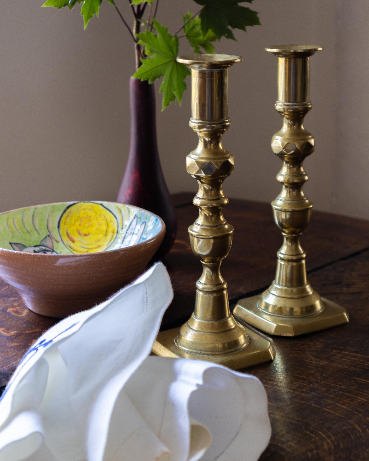 Two brass candlesticks on a wooden surface with a ceramic bowl and decorative vase in the background.