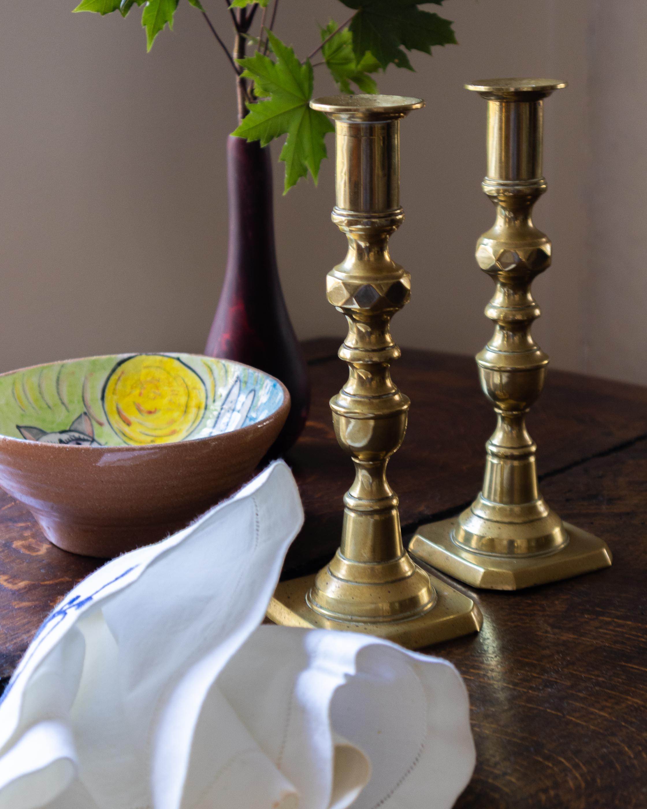 Two brass candlesticks on a wooden surface with a ceramic bowl and decorative vase in the background.