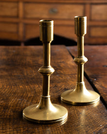 Two brass candle holders on a wooden surface with a wooden drawer in the background.
