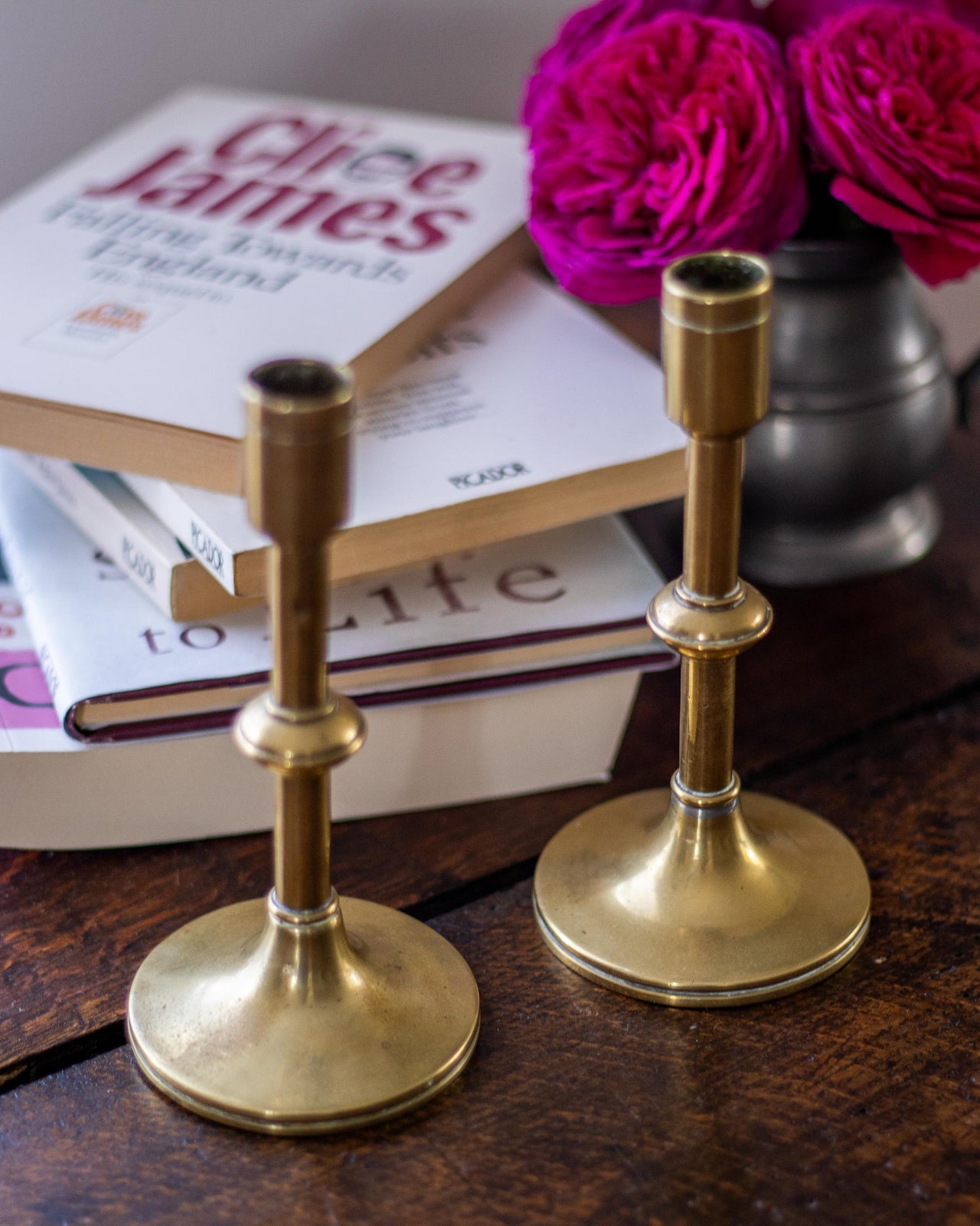 Two brass candle holders on a wooden surface with books and flowers in the background.