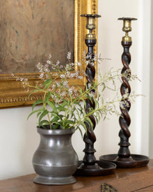 Decorative plant in a pot with two wooden candlesticks on a surface, framed by a painting.