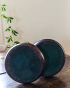 Two dark bases of candlesticks on a wooden surface with a plant in the background
