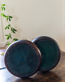 Two dark bases of candlesticks on a wooden surface with a plant in the background