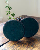 Two dark green circular bases of candlesticks on a wooden surface with a plant in the background.