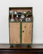 Vintage wooden toy cabinet with green trim filled with metallic kitchenware on a wooden surface.