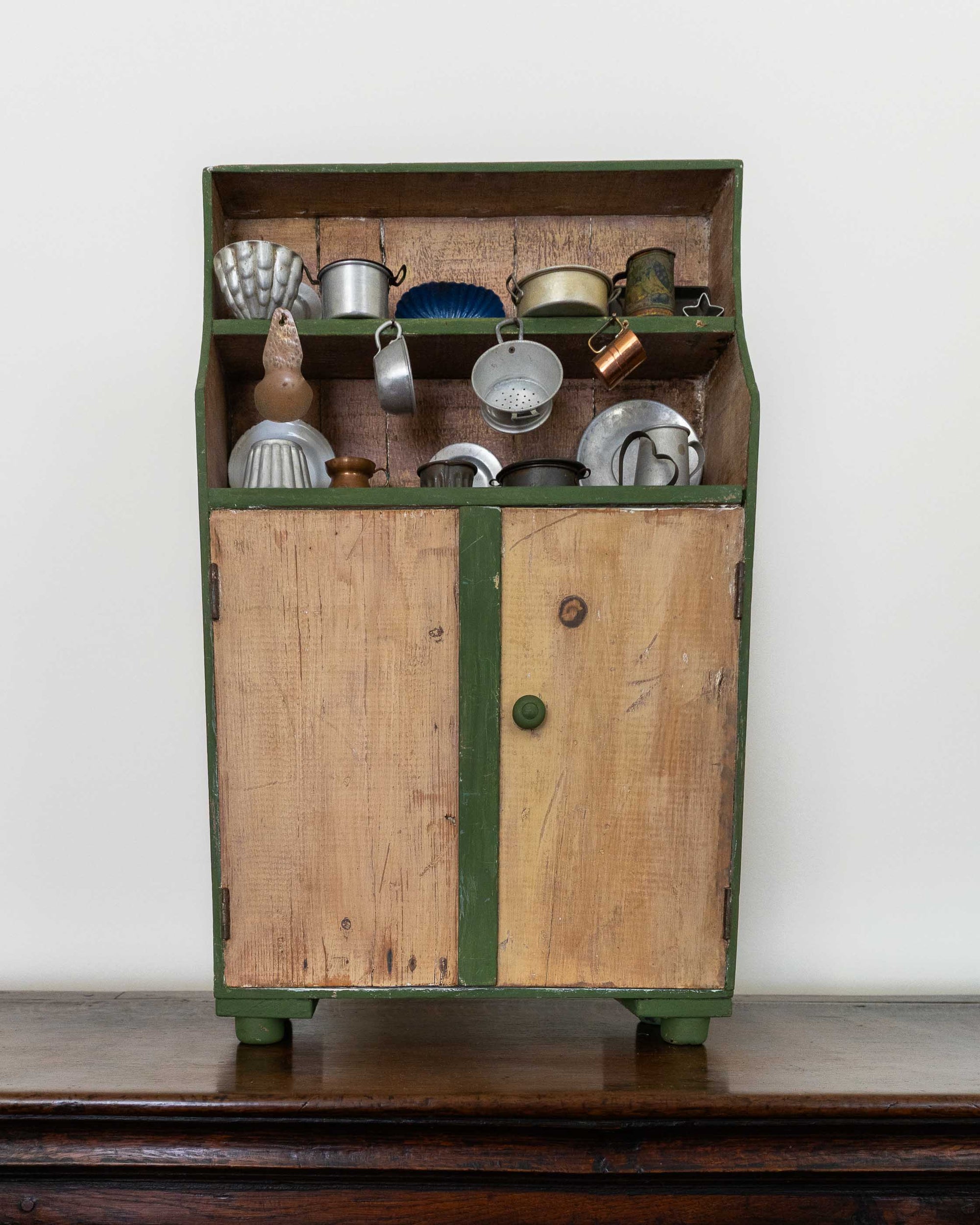 Vintage wooden toy cabinet with green trim filled with metallic kitchenware on a wooden surface.