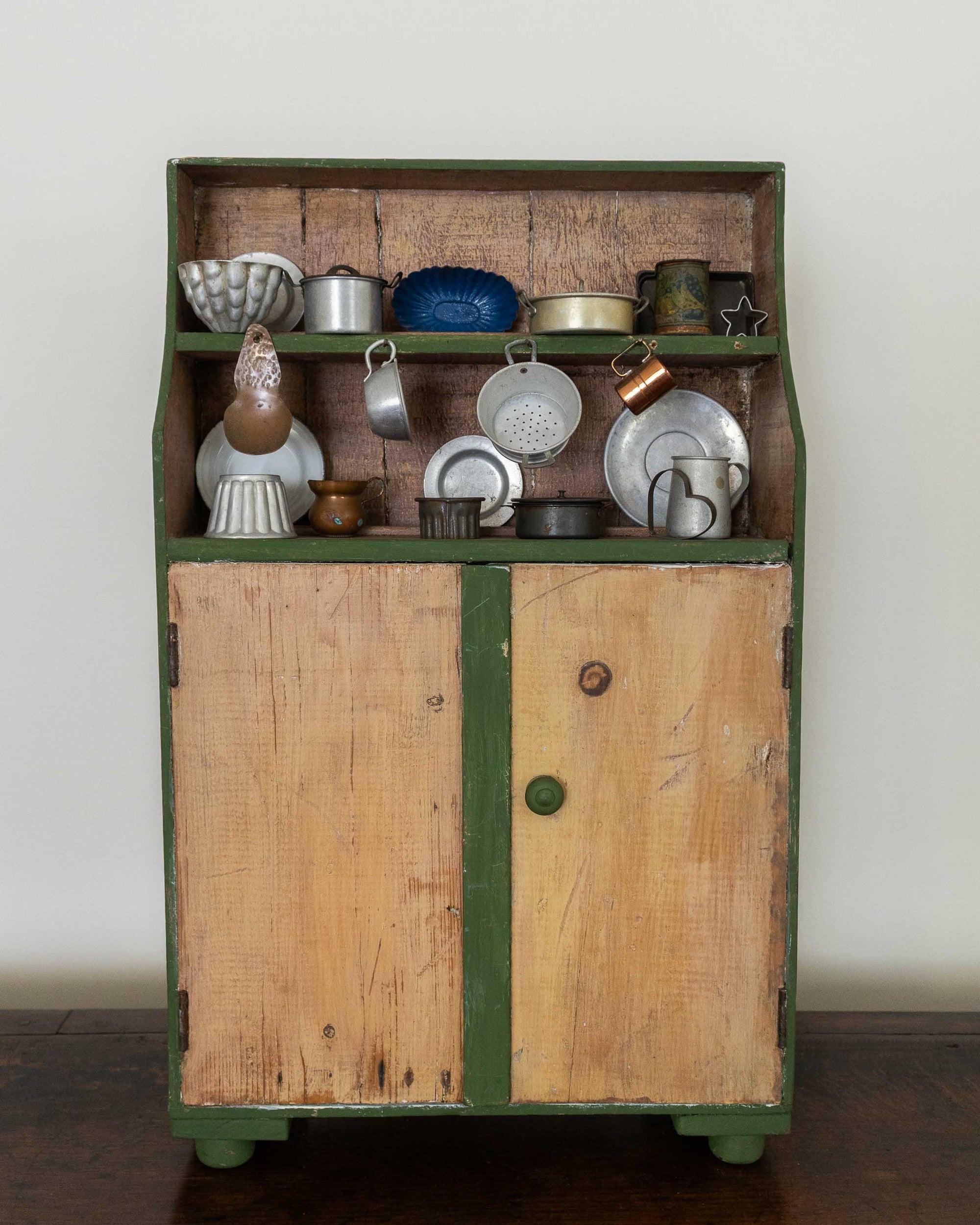 Wooden toy cabinet with green trim displaying various ceramic items against a white wall.