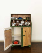 Wooden toy cabinet with open door displaying miniature kitchen items on a white background