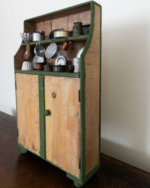Wooden toy cabinet with green trim and various kitchen items on a white wall background