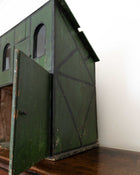 Green wooden stables with a door open on a wooden floor against a white wall.
