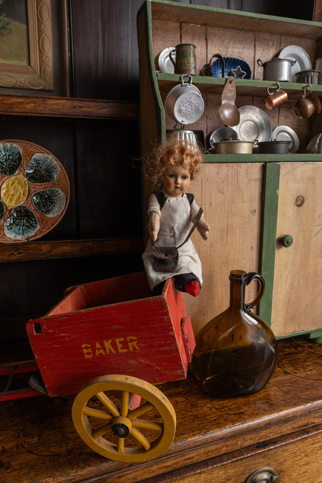 Doll with red cart and  kitchen dresser with kitchen items on a wooden shelf