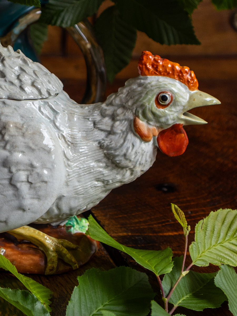 Decorative chicken figurine with a red comb on a wooden surface with green leaves.