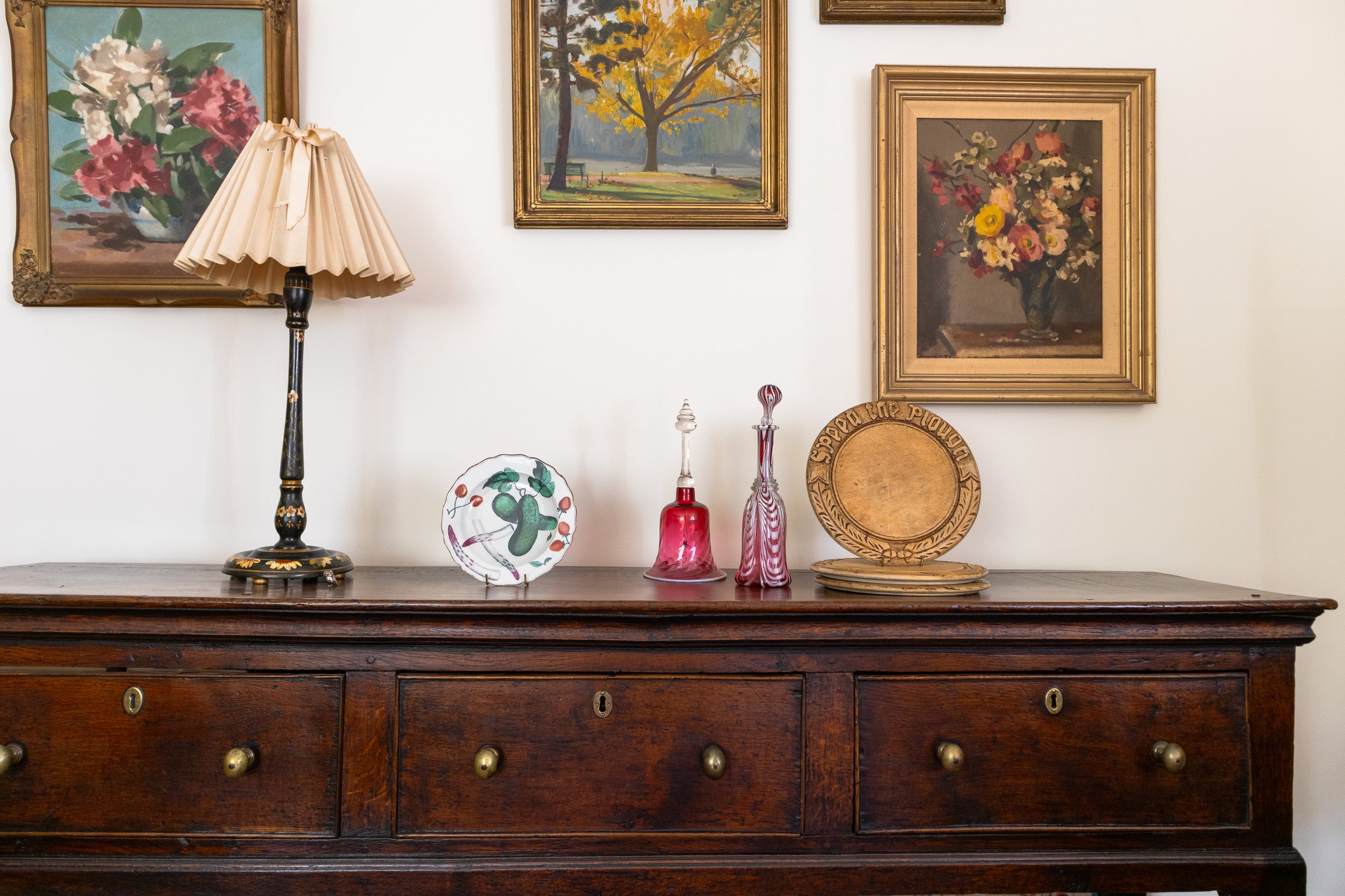 Wooden dresser with decorative antiques and framed paintings on a wall.