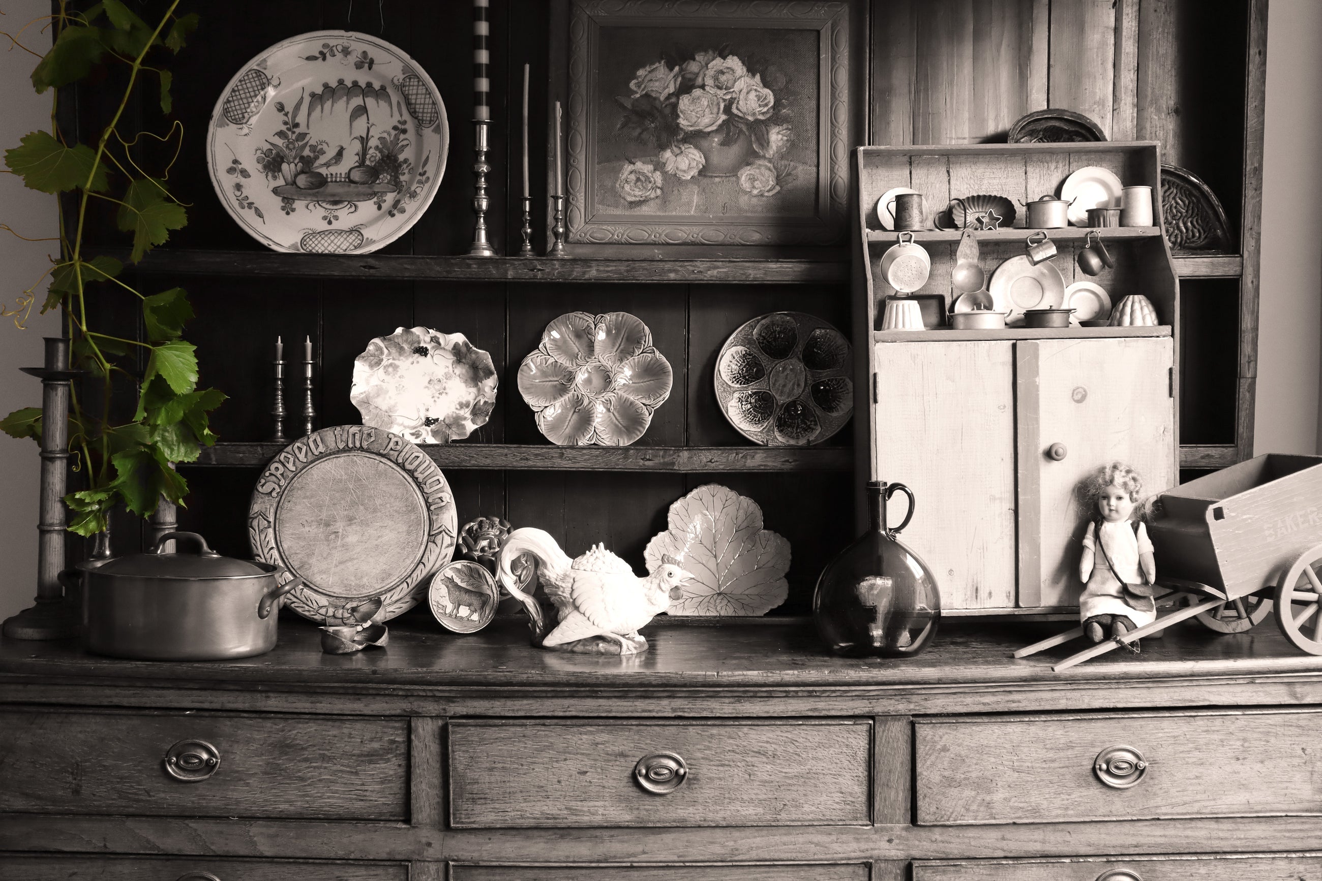 Dresser with a collection of ceramics plates, doll and cart, painting and green leaves on a black and white background