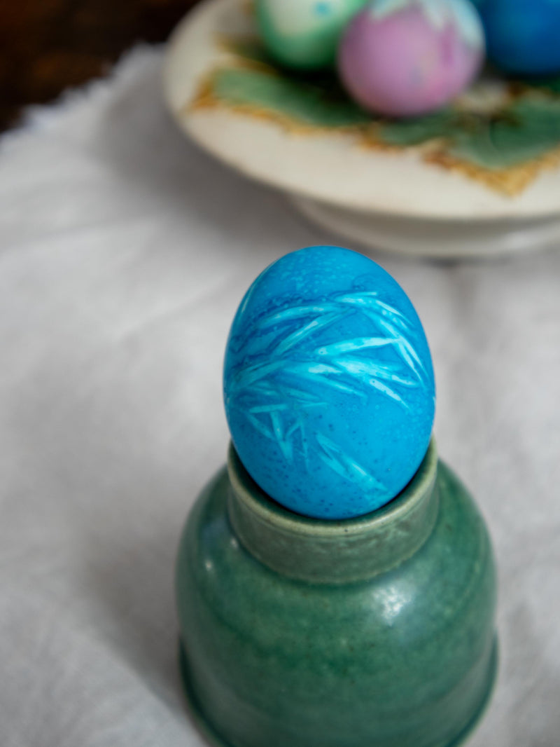 Blue dyed egg on a green ceramic stand with blurred Easter eggs in the background.