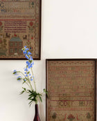 Framed cross-stitched sampler artworks on a wall with a vase of blue flowers.