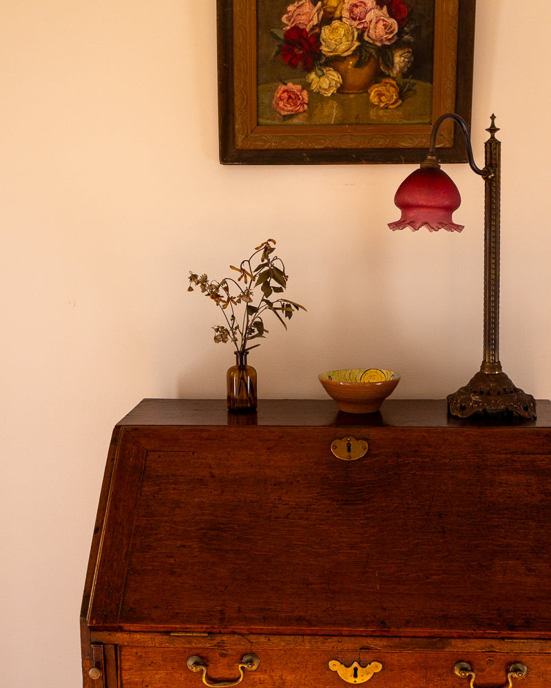 Antique desk with a lamp, bowl, and vase on top, with a floral painting in the background.