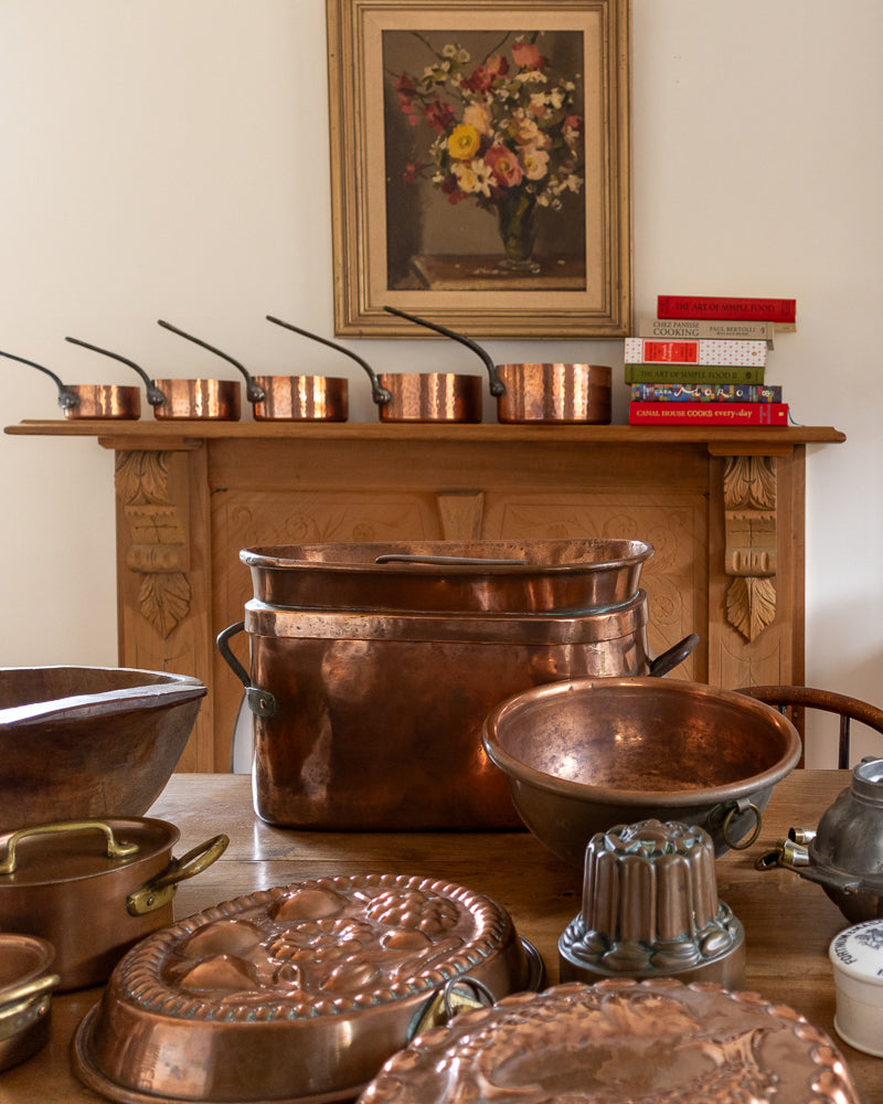 Collection of copper cookware on a wooden table with a floral painting in the background.
