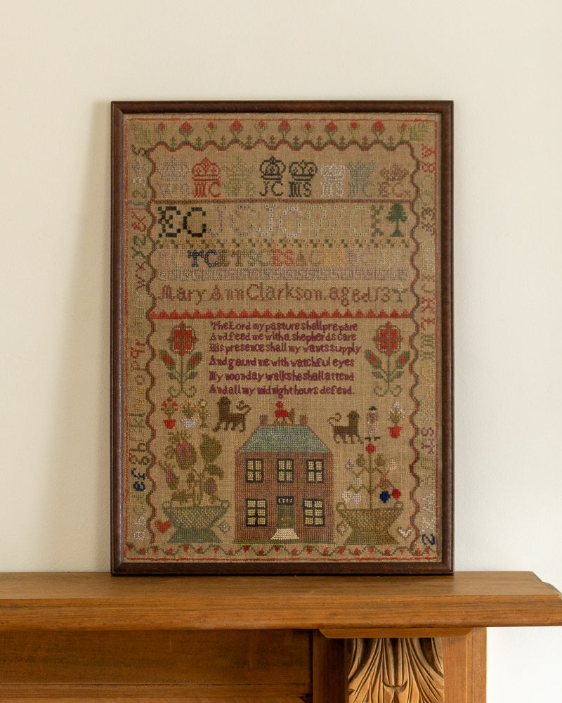 Framed needlepoint sampler with decorative elements and text on a wooden surface.