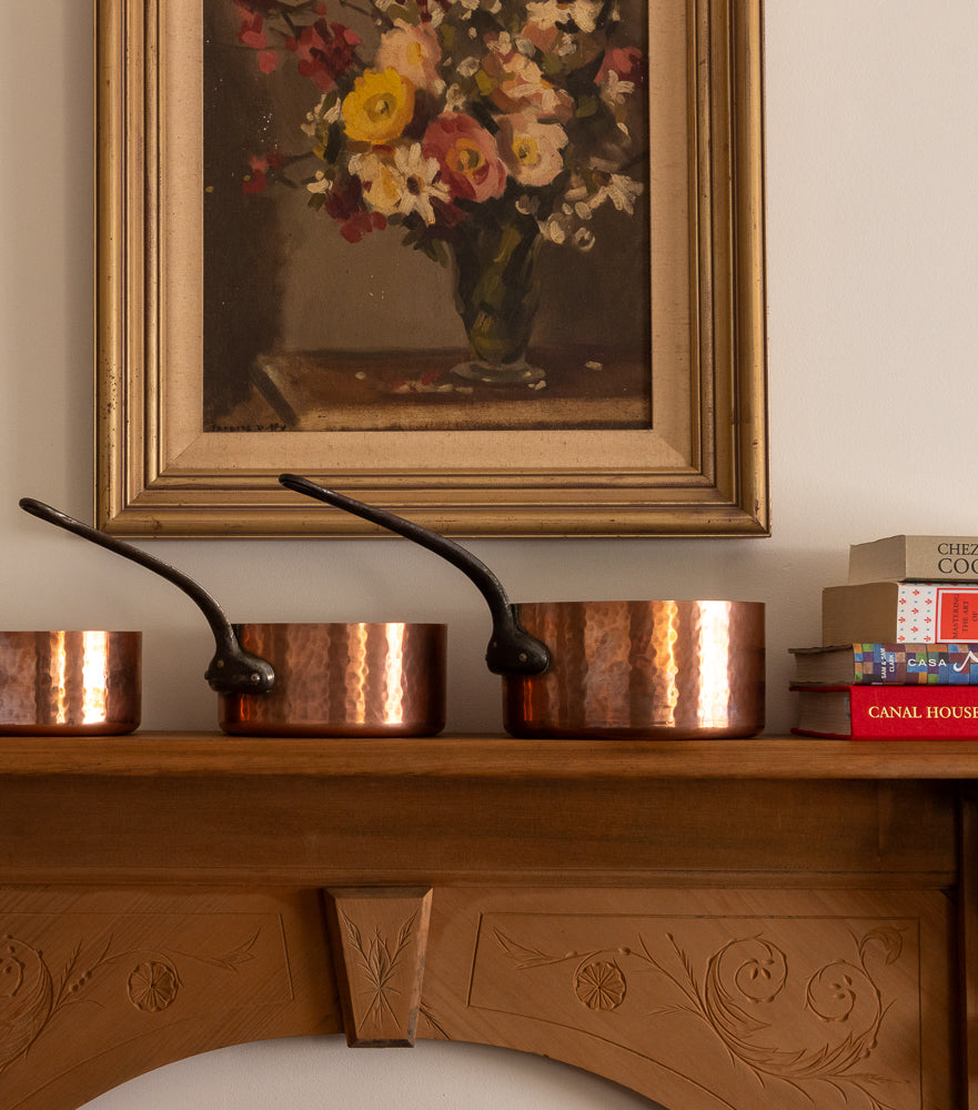 Copper pots on a wooden surface with a floral painting in the background