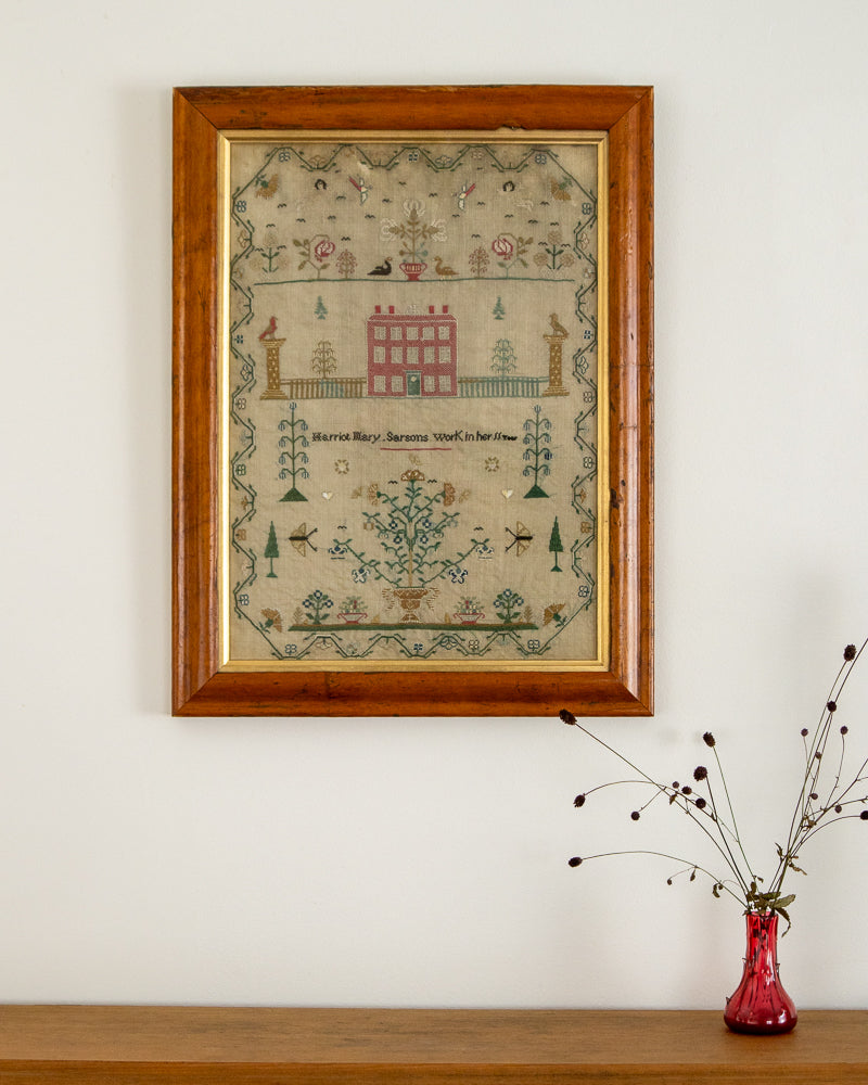 Framed sampler on a wall with a vase of dried flowers below