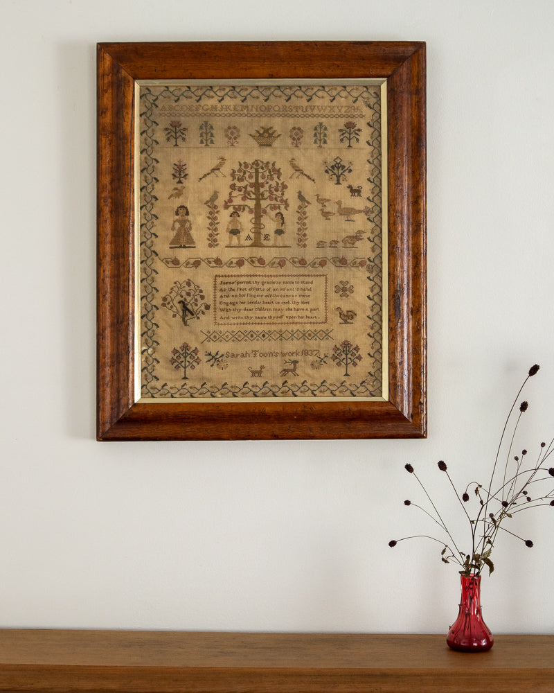 Framed sampler art on a wall with a vase of dried flowers below